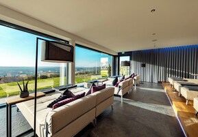 Quarto de hotel moderno com uma janela ampla que oferece uma vista panorâmica, um sofá com almofadas, uma TV de tela plana e uma área de jantar com mesa e cadeiras.