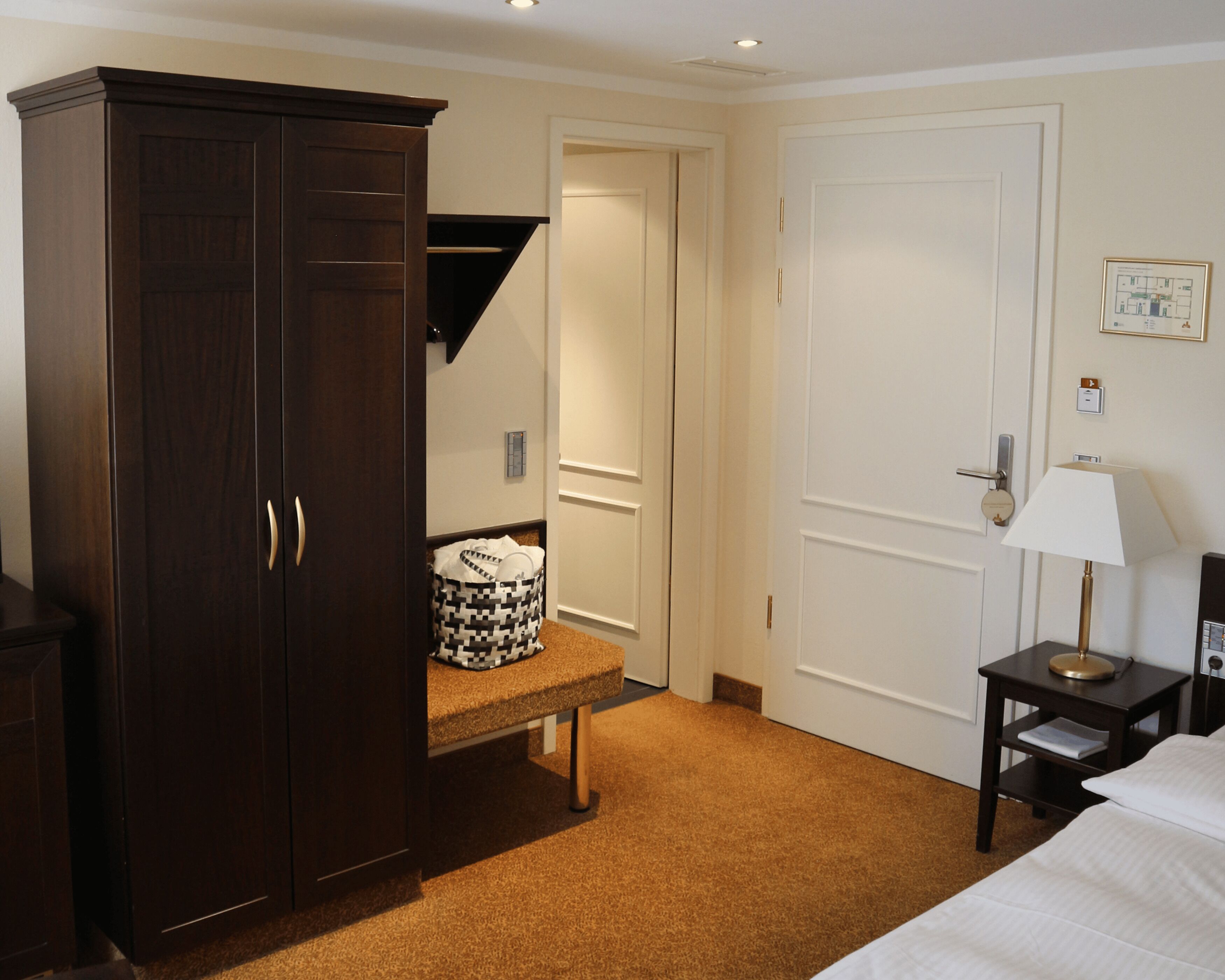 A hotel room with a large wardrobe, a bedside table with a lamp, and a carpeted floor.