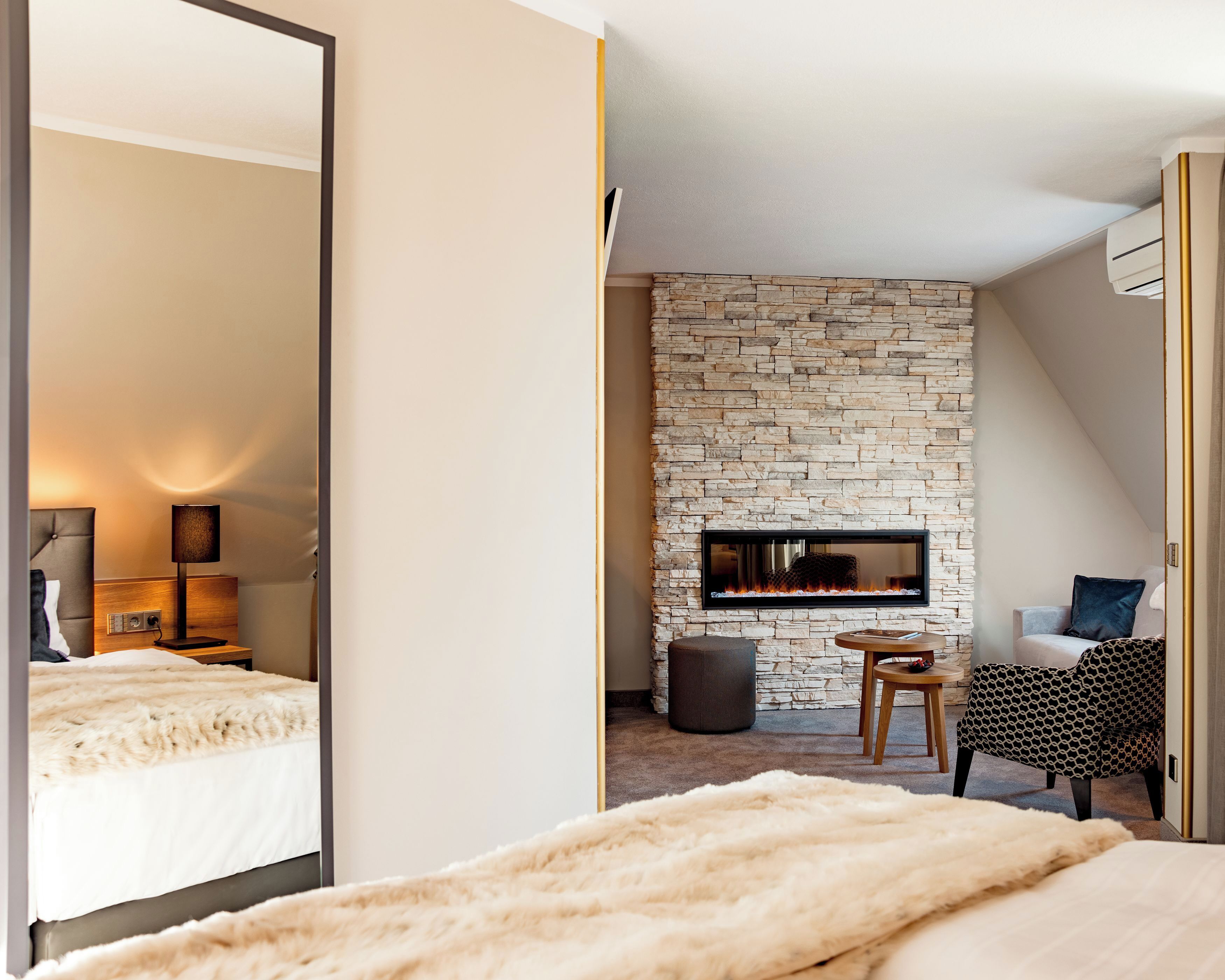 Une chambre d’hôtel moderne avec une cheminée en pierre, un grand miroir, un lit recouvert d’une couverture blanche moelleuse et un coin salon avec un fauteuil à motifs.
