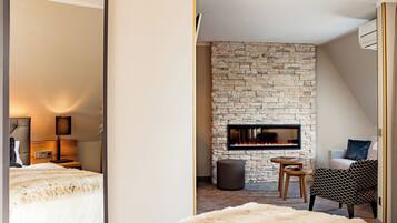 Une chambre d’hôtel moderne avec une cheminée en pierre, un grand miroir, un lit recouvert d’une couverture blanche moelleuse et un coin salon avec un fauteuil à motifs.