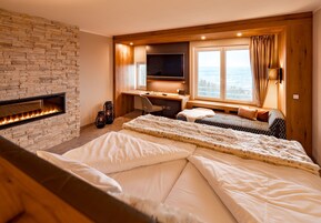 Quarto com lareira de pedra, escrivaninha com computador, cama com roupa de cama branca e uma janela grande com vista.