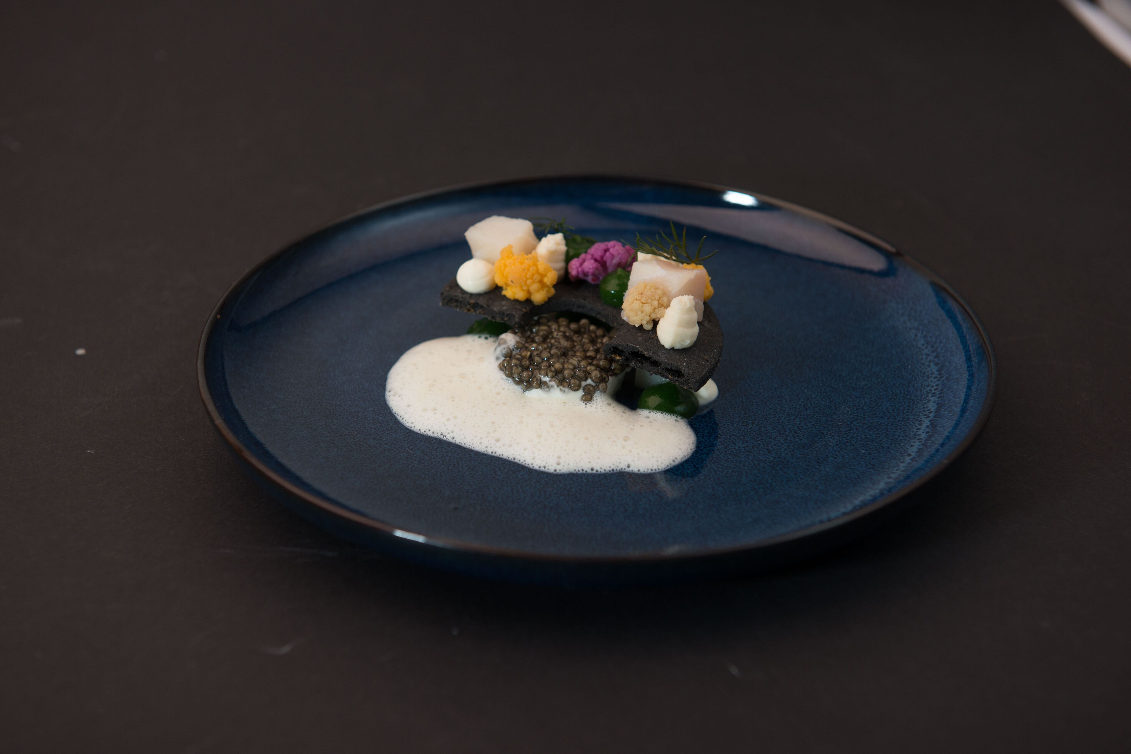 A dark blue plate with a white creamy substance, a piece of fish, and various garnishes on a dark surface.