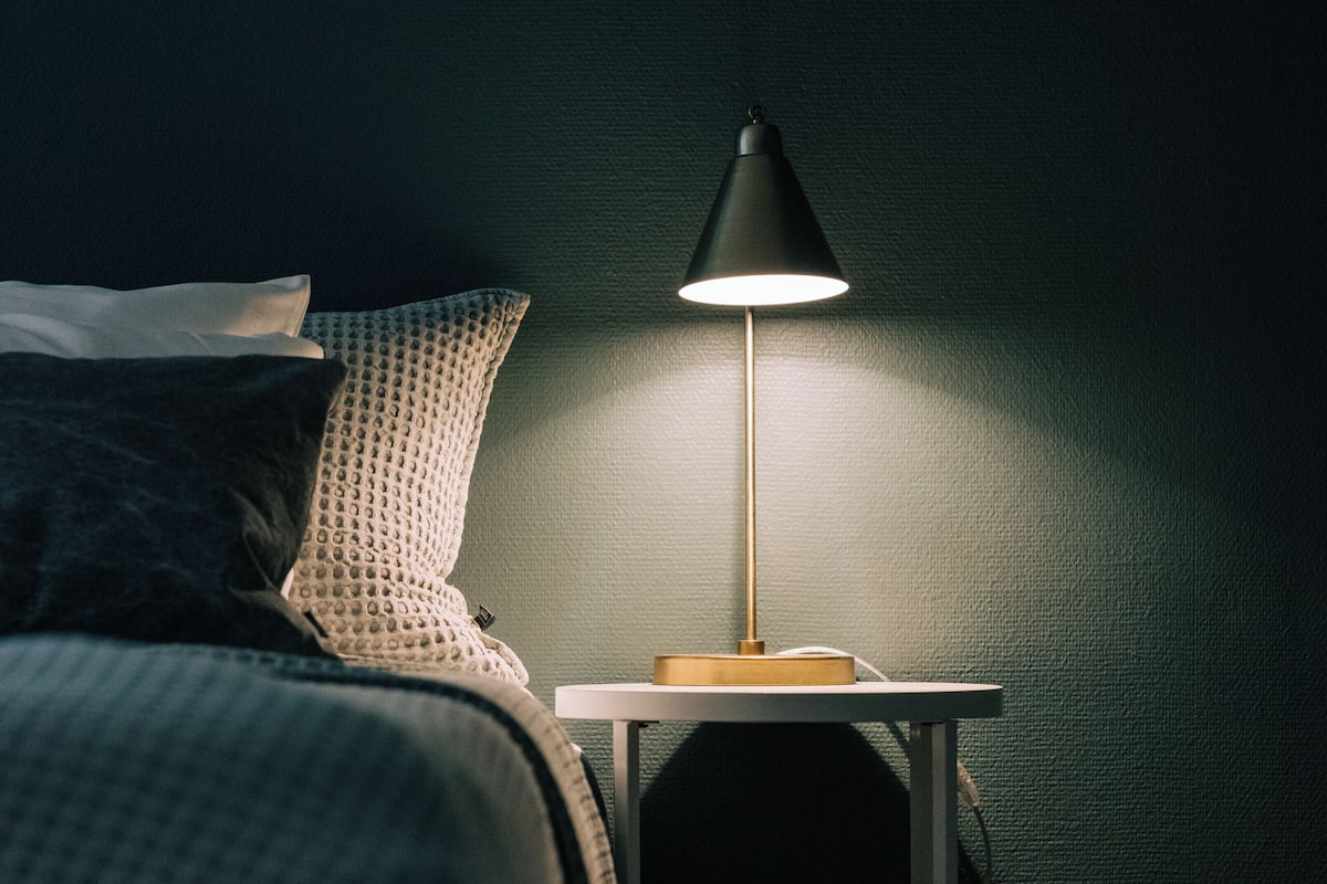 A neatly made bed with a checkered bedspread and pillows, a small white nightstand with a lamp, and a wooden floor.