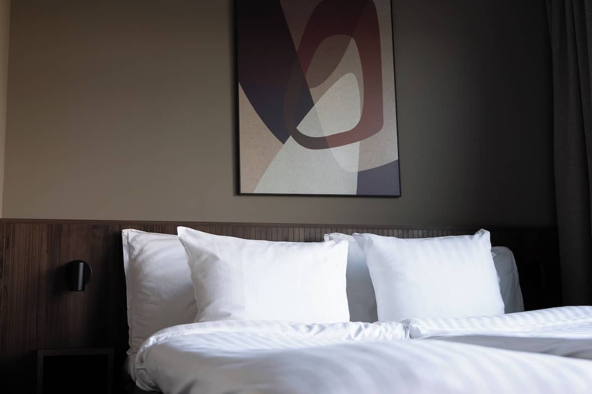 A hotel room with a neatly made bed, a wall-mounted light, and a large abstract artwork above the bed.