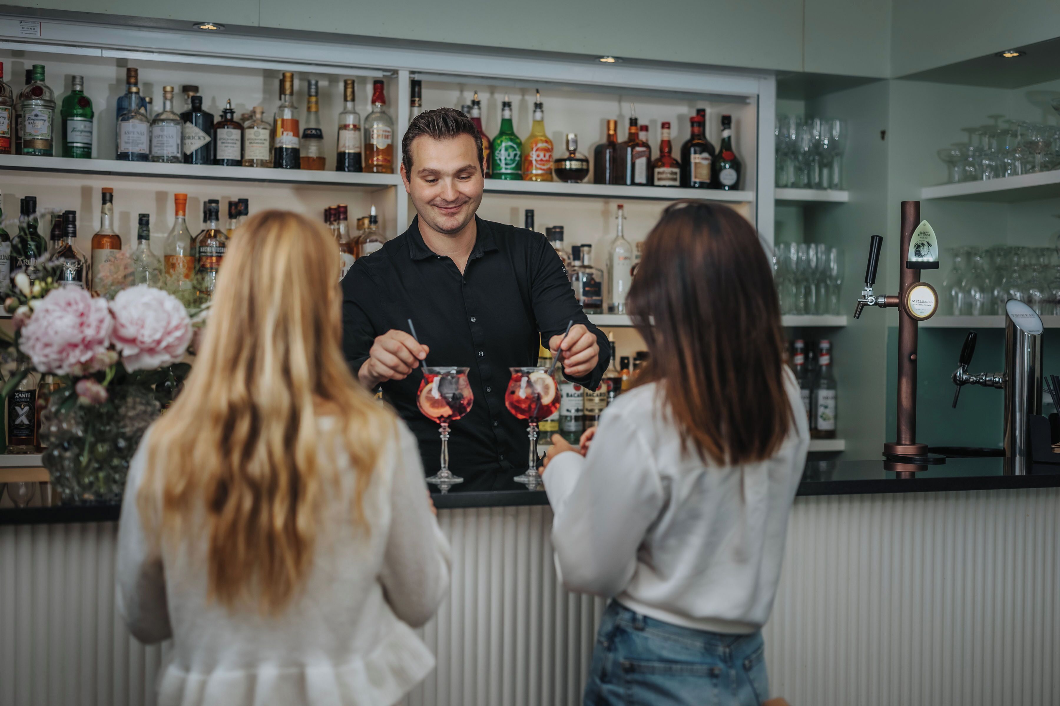 Een bar met flessenkast vol drank, een toog en iemand die drankjes uitserveert.