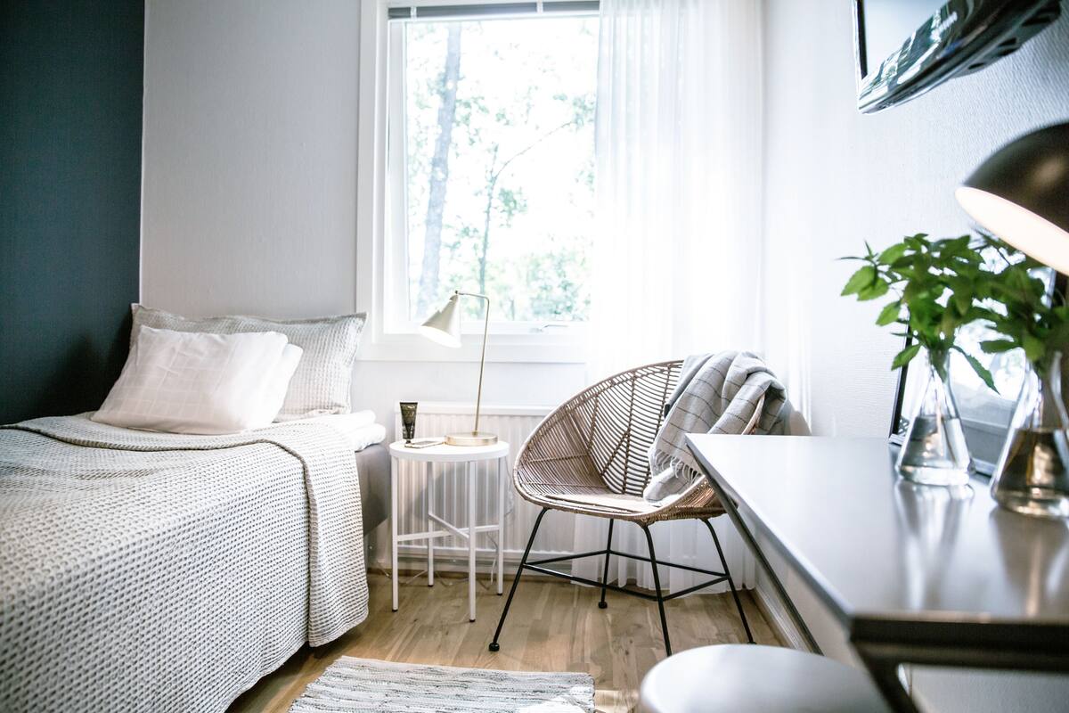 A bedroom with a bed, a small round table, a chair, a lamp, and a window with a view of trees.