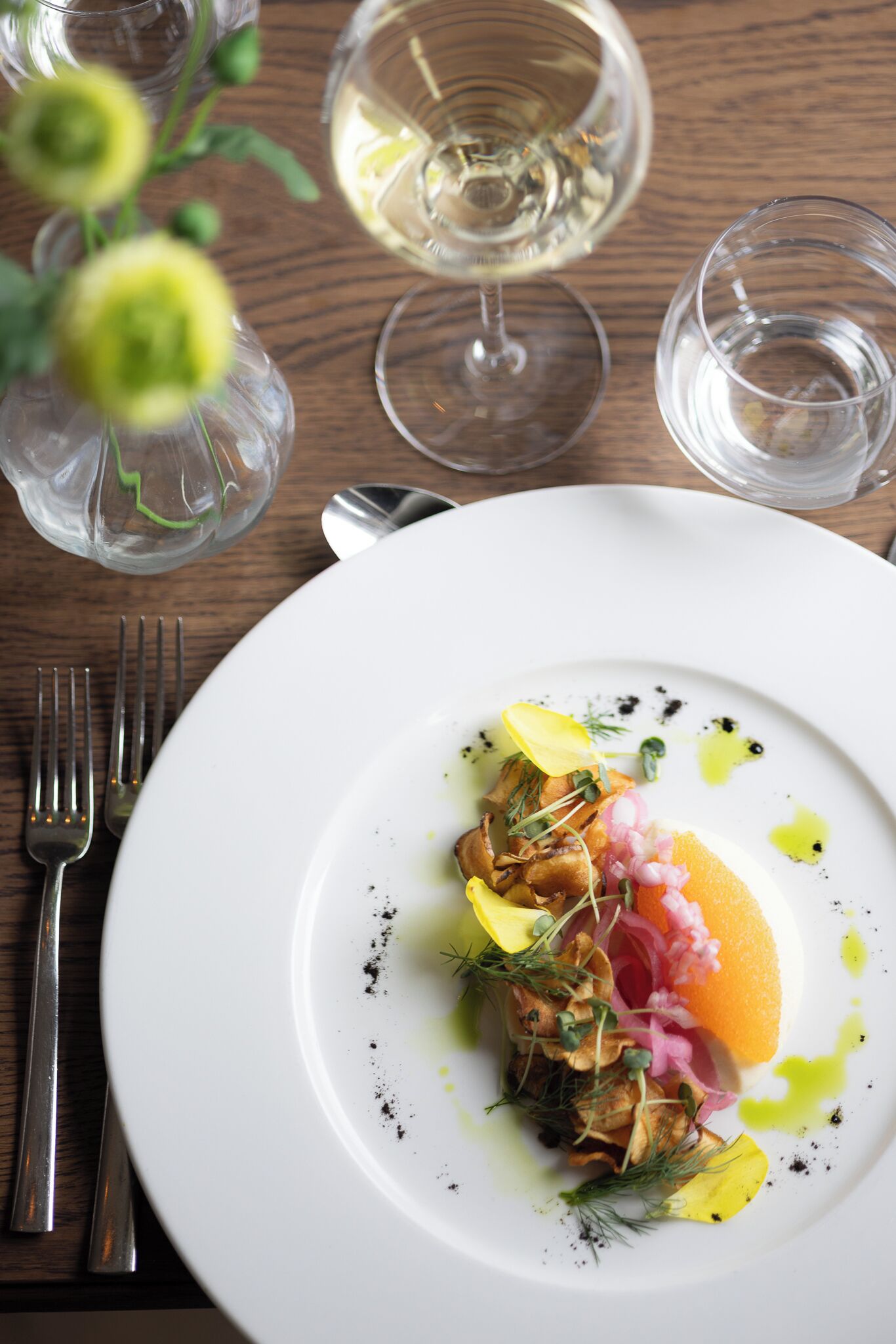 A white plate with a dish, a fork, a spoon, and two glasses on a wooden table.