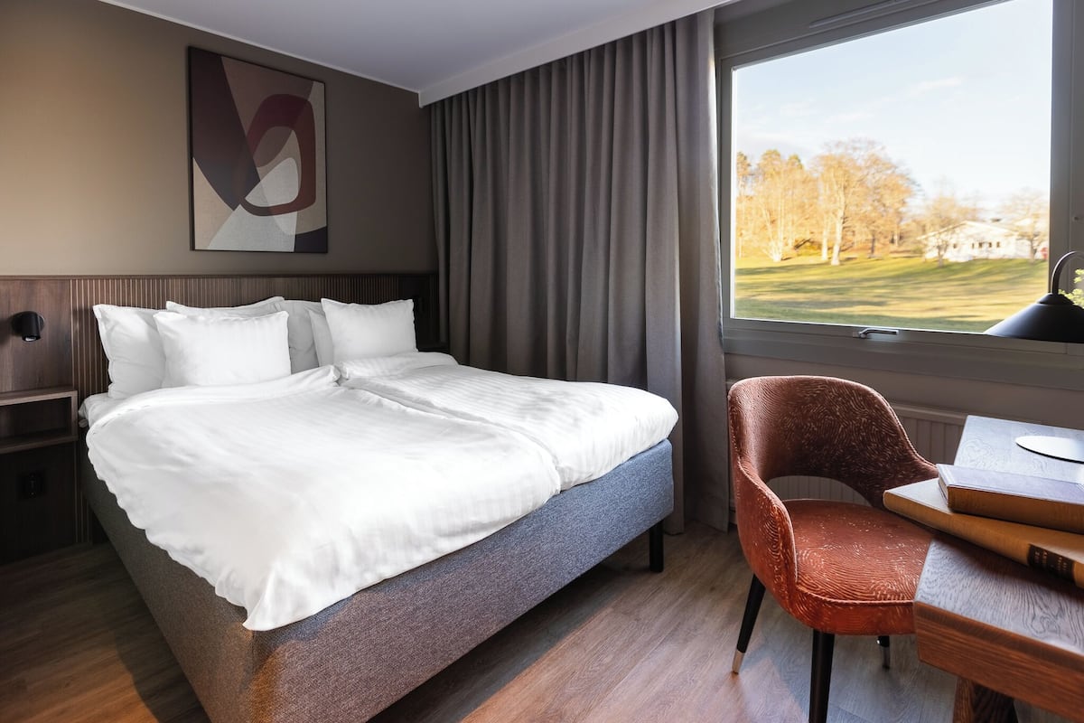 A hotel room with a bed, a chair, a table, a window with a view of trees and a building, and a wall-mounted artwork.