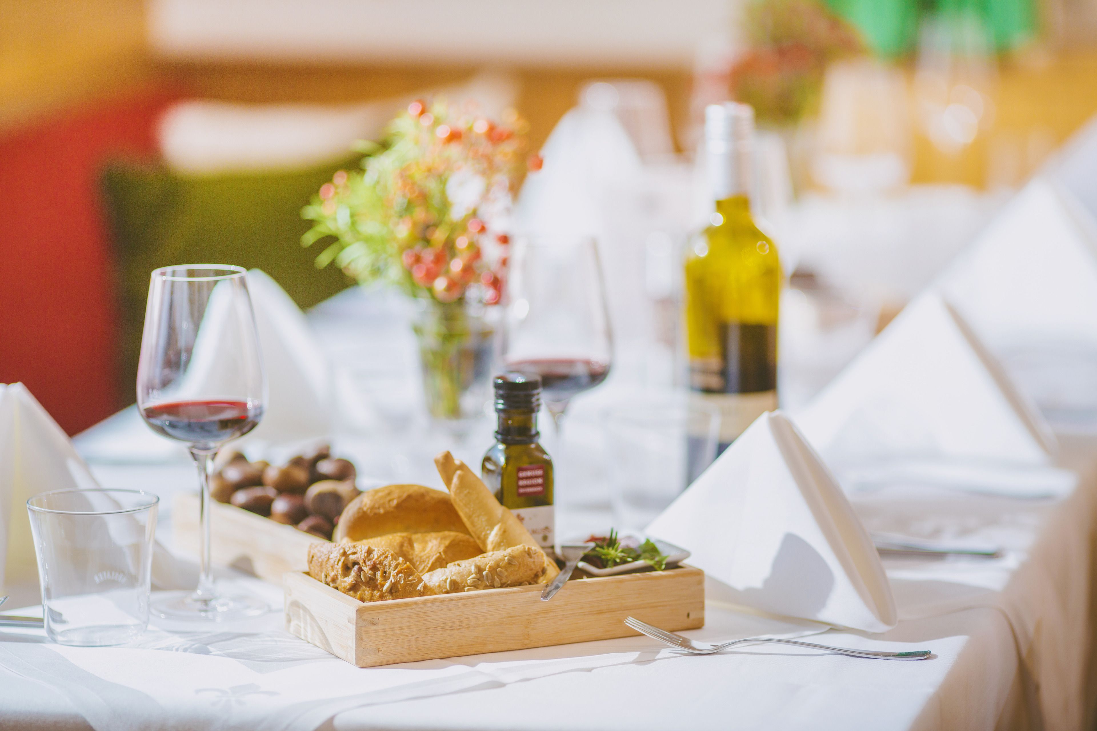 A dining table with a wooden serving tray, glasses of red wine, a bottle of olive oil, and a small bowl of nuts.