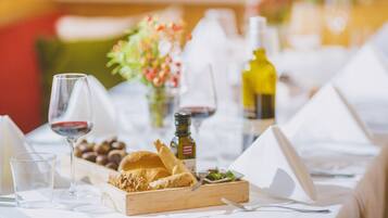 A dining table with a wooden serving tray, glasses of red wine, a bottle of olive oil, and a small bowl of nuts.