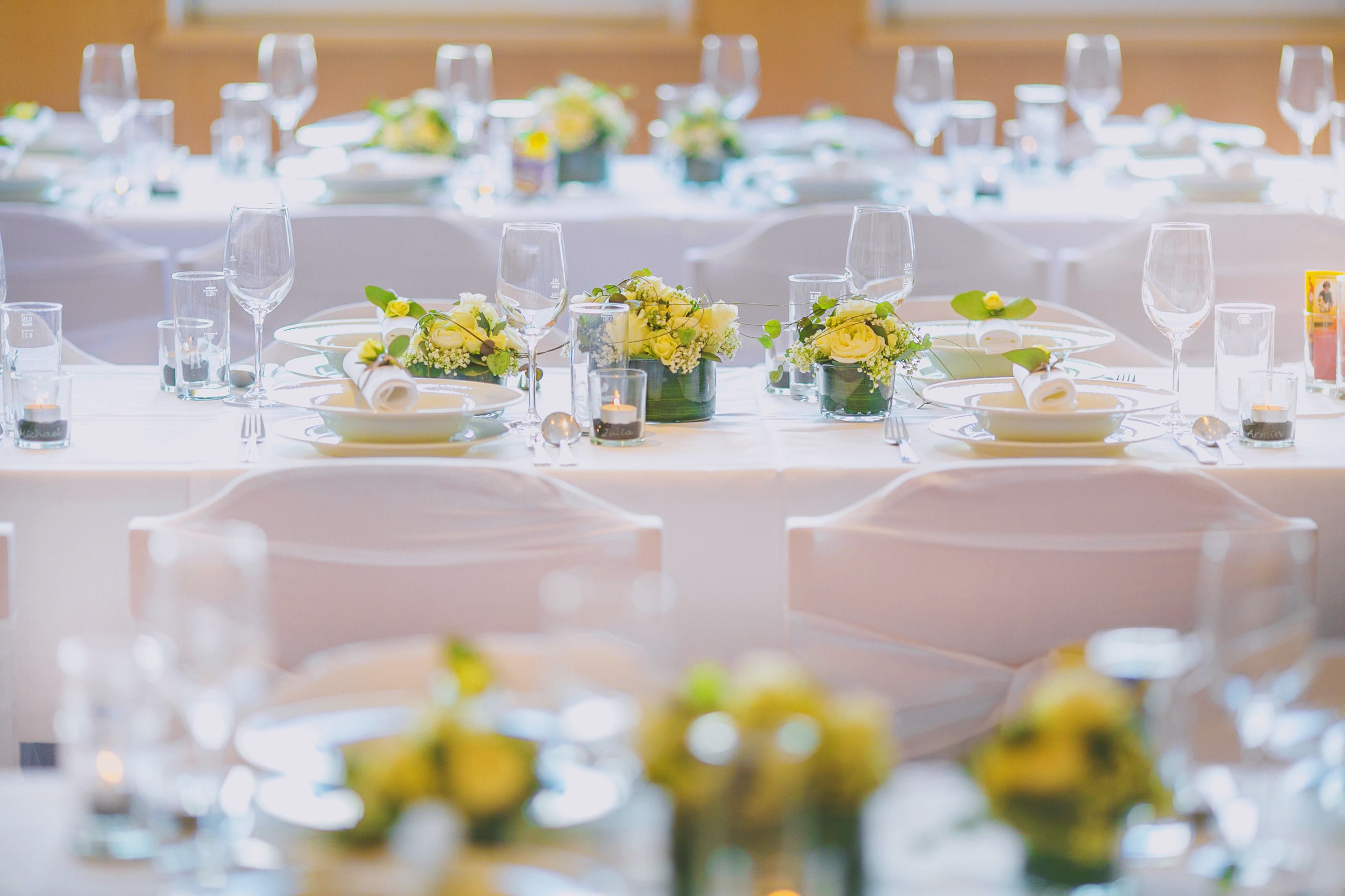 Een balzaal met ronde tafels gedekt voor een formeel evenement, elk met een bloemstuk van gele bloemen en helder glaswerk.