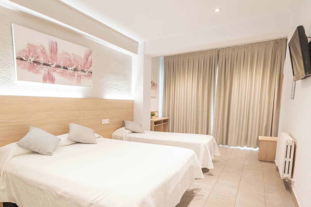 A hotel room with two beds, a large window with curtains, a flat-screen TV, and a wall-mounted artwork.