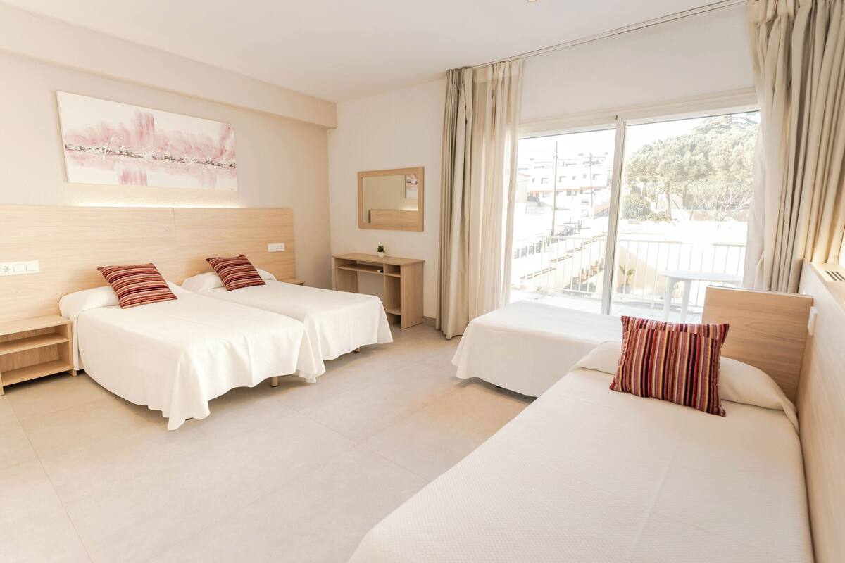 A hotel room with two single beds, a small desk, a mirror, and a large window with curtains.