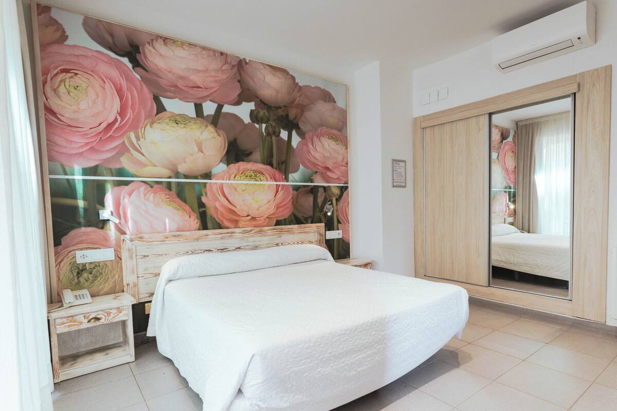 A neatly made bed in a room with a large floral mural on the wall, a wooden wardrobe, and a mirror reflecting part of the room.