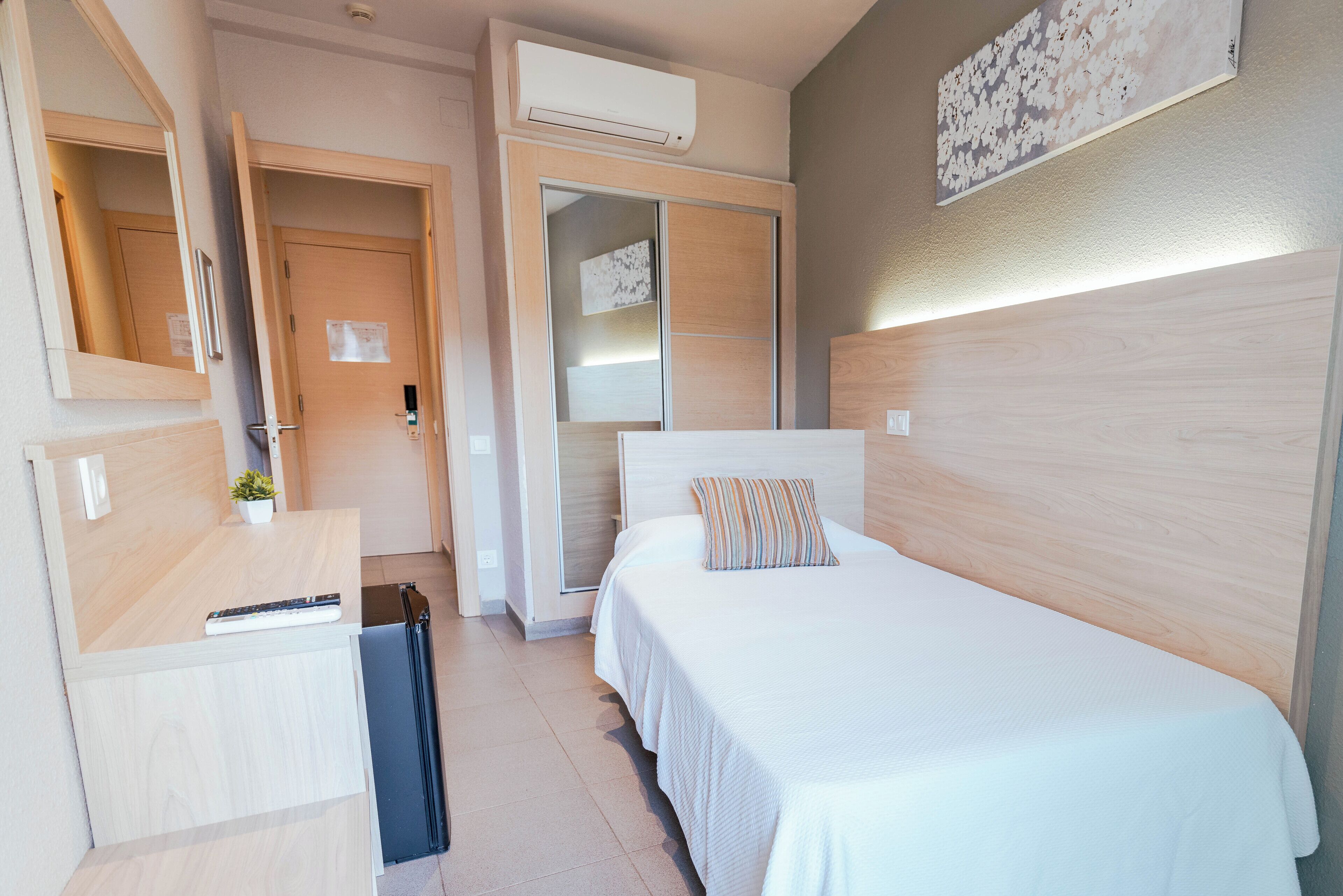 A hotel room with a bed, a desk, a mirror, a wardrobe, and an air conditioning unit.