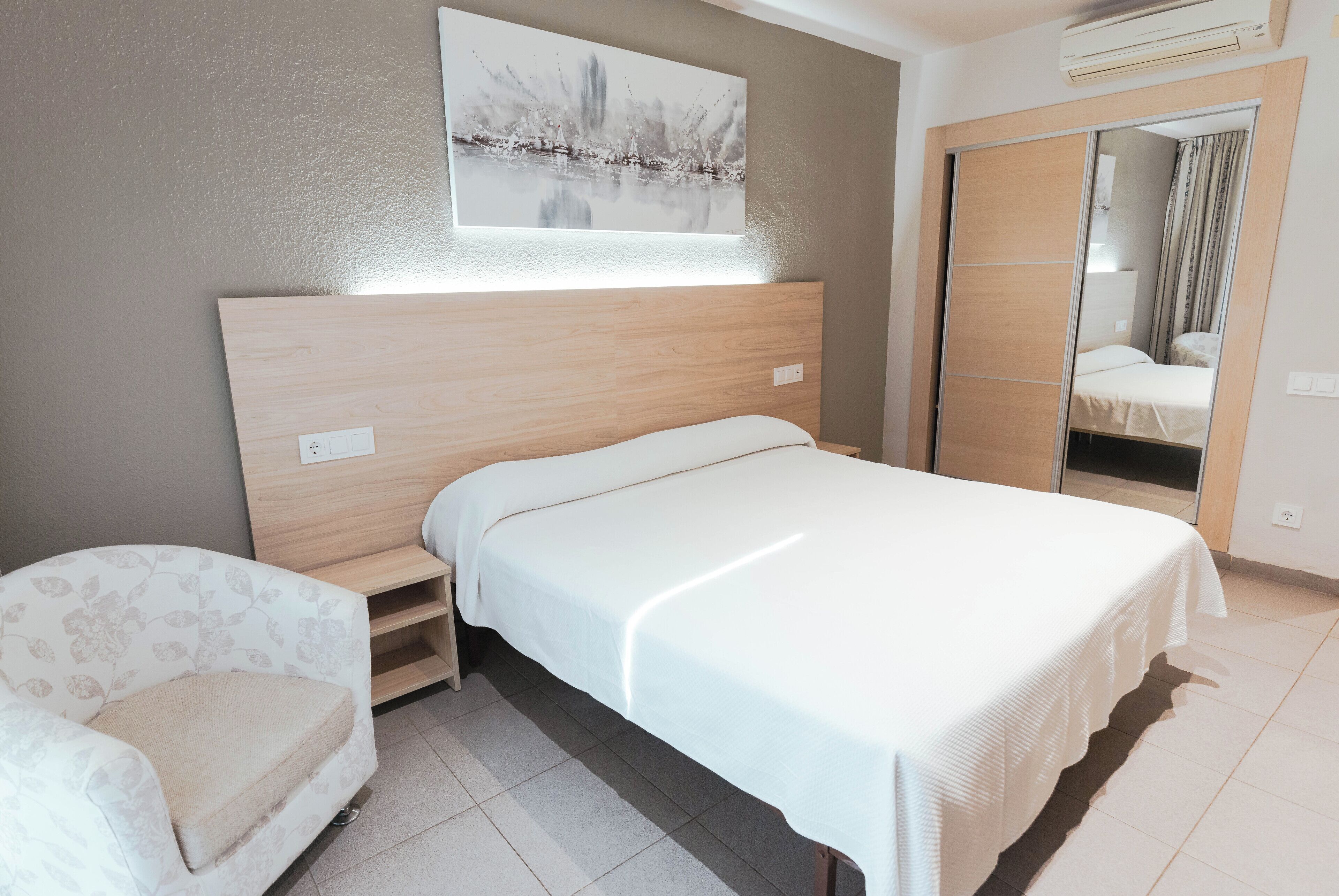 Un dormitorio con cama, una silla, una mesita de noche, un espejo y un cuadro en la pared.