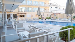 Zona junto a la piscina con sillas de descanso y sombrillas, un edificio con balcones y un cartel.