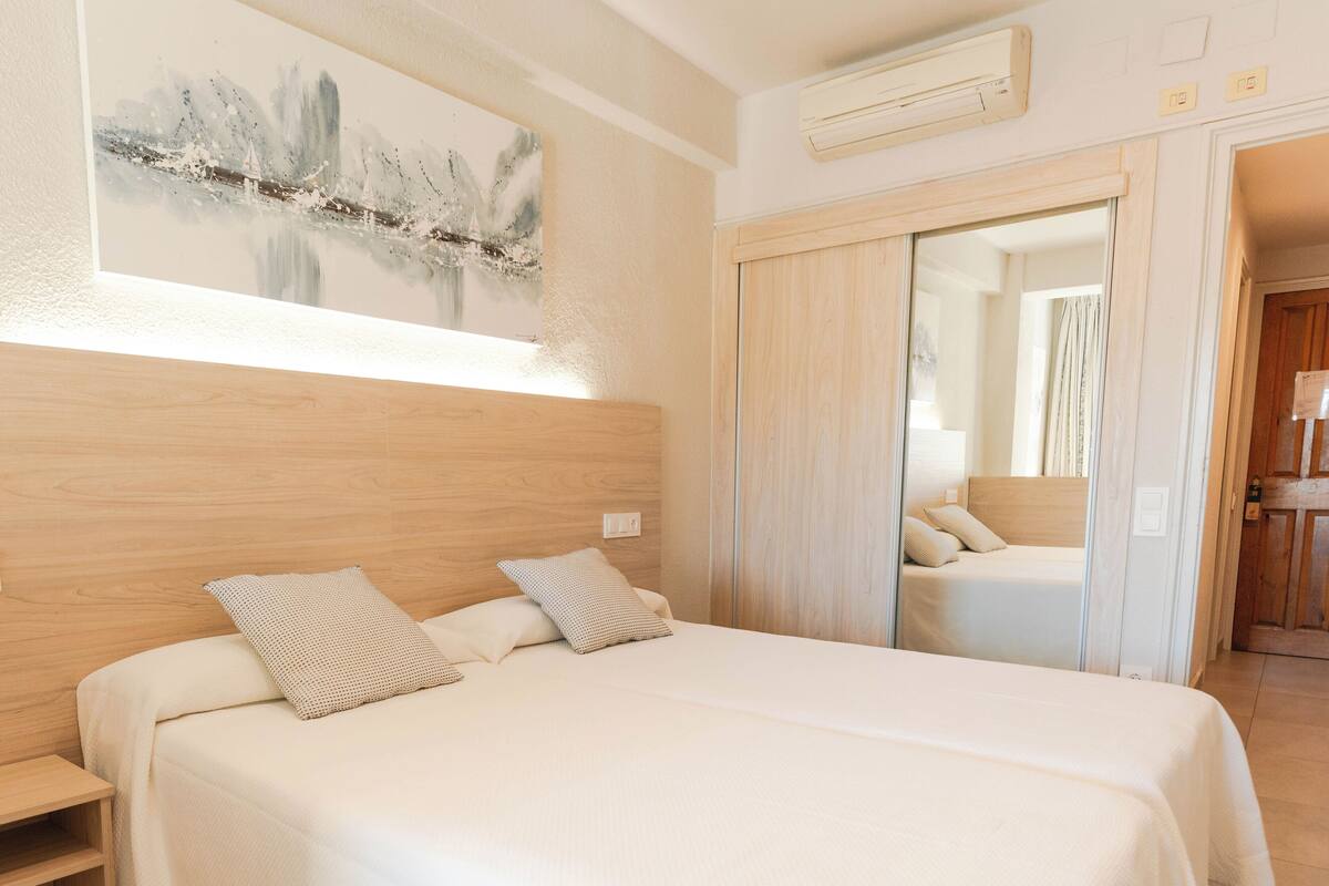 A hotel room with two beds, a wooden headboard, a painting on the wall, and a mirror reflecting the room.