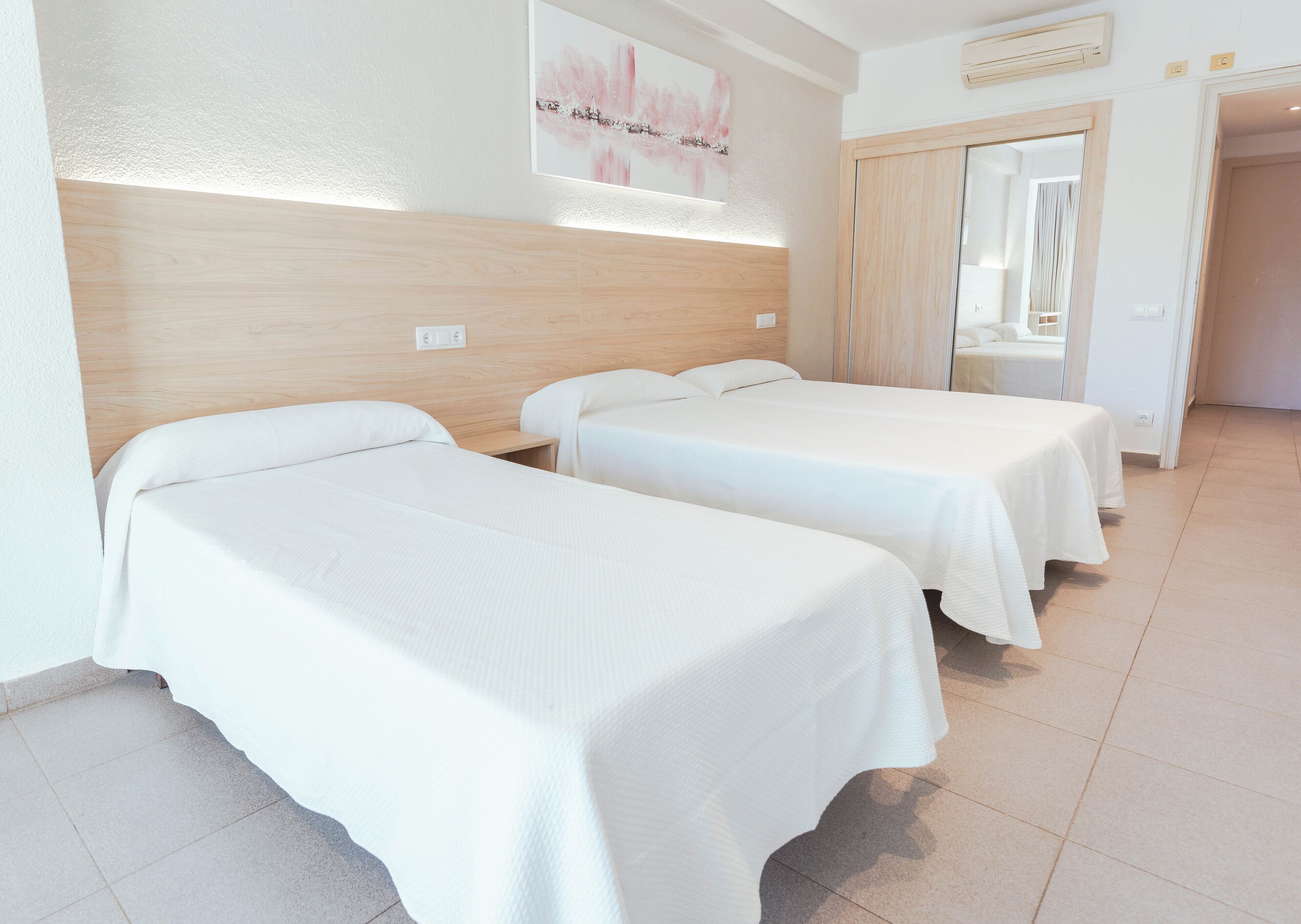 Habitación de hotel con dos camas individuales, cabecero de madera, pared blanca con un cuadro y suelo de baldosas.