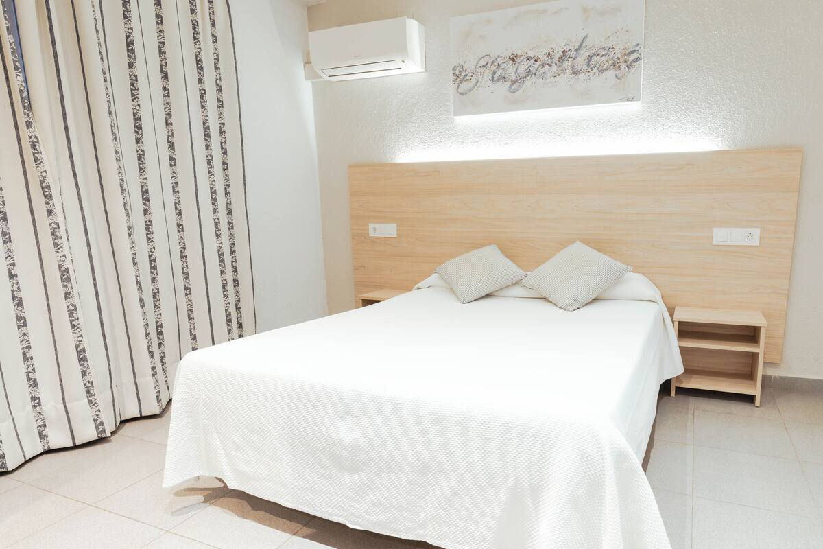 A neatly made bed with a white bedspread and two pillows, a wooden headboard, a small wooden nightstand, and a window with a view of the sky.