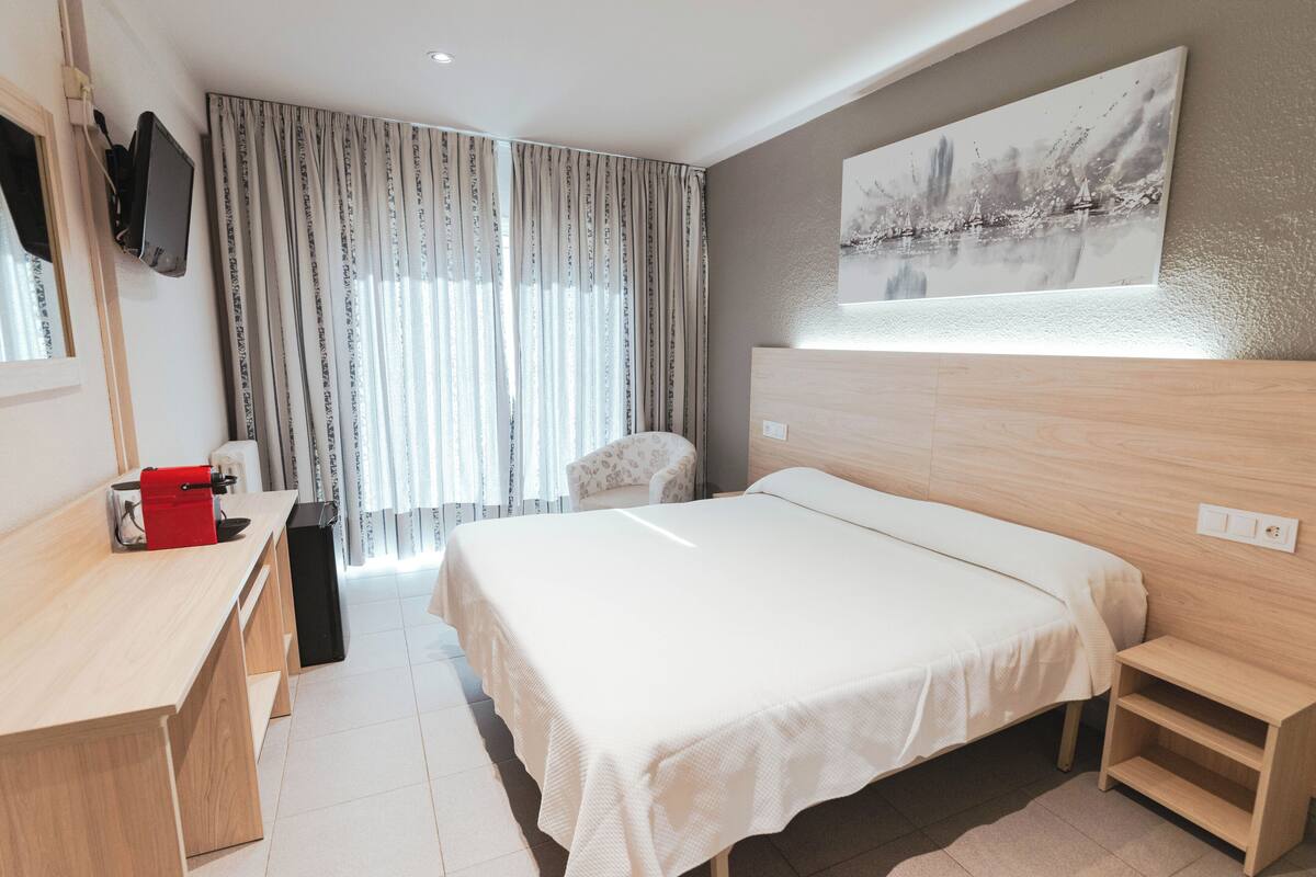 A modern hotel room with a large bed, a desk, and a chair. There is a window with curtains, a painting on the wall, and a TV mounted on the wall.