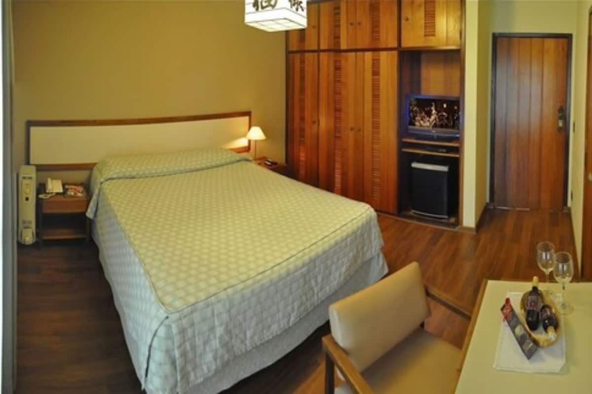 Ein Hotelzimmer mit einem großen Bett, einem Fernseher, einem Holzschrank, einem kleinen Tisch mit Flasche und Gläsern sowie einem Sessel.