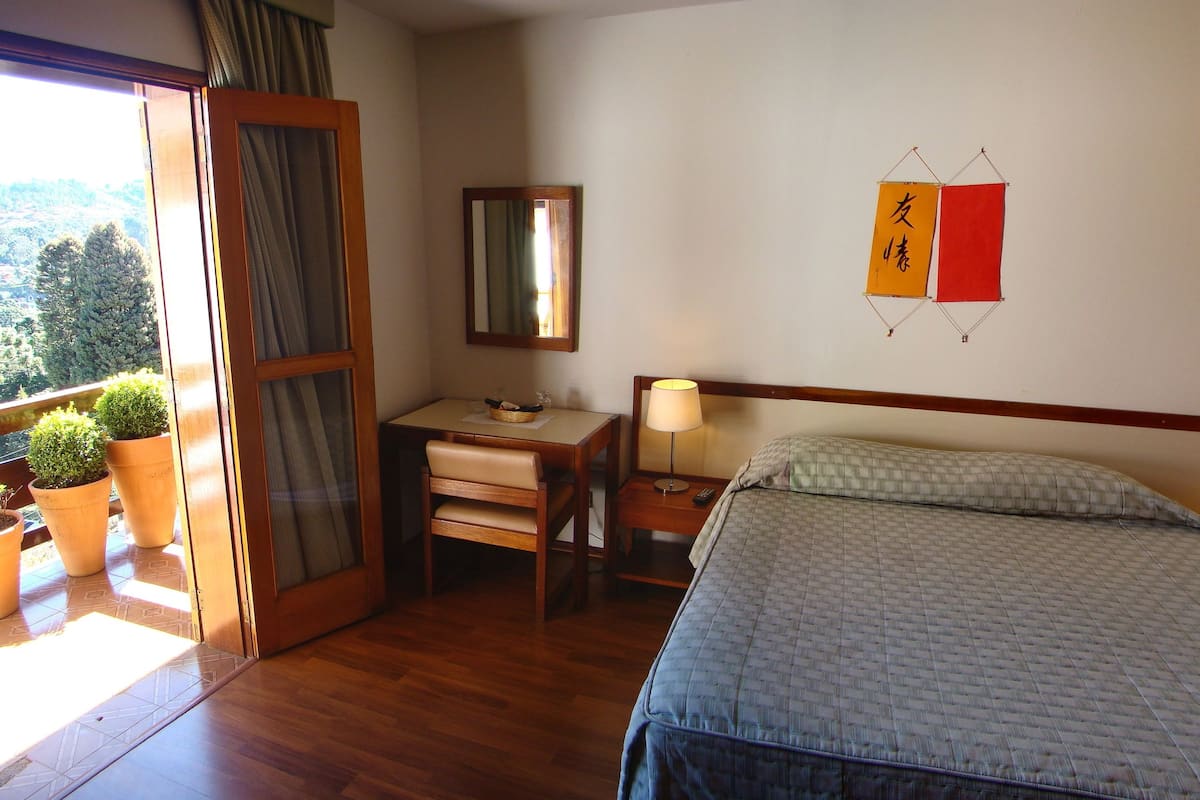 Ein Hotelzimmer mit einem Bett, Nachttischen mit Lampen, einem Schreibtisch, einem Stuhl, einem Spiegel und Blick ins Freie.
