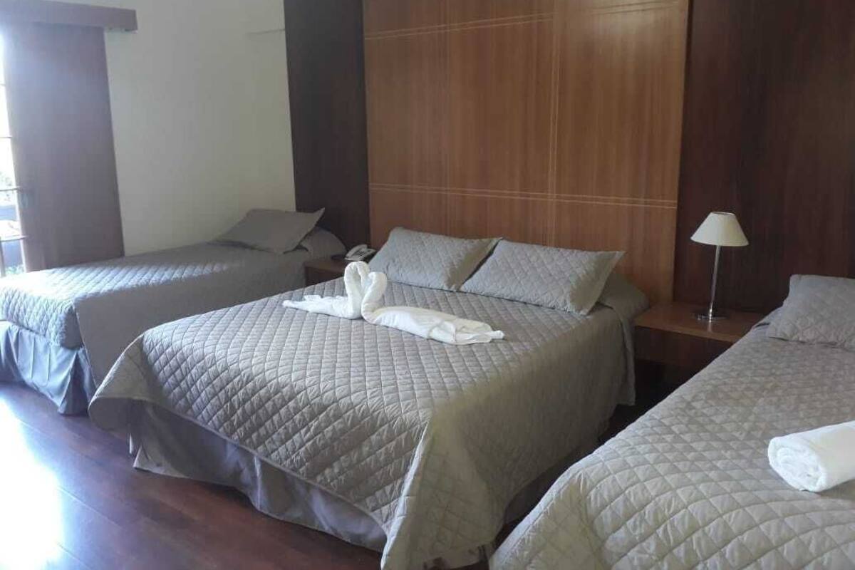 Ein Hotelzimmer mit drei Betten, jeweils mit gesteppter Bettdecke und Kissen. Es gibt ein hölzernes Kopfteil, eine Nachttischlampe und ein Fenster mit Vorhängen.