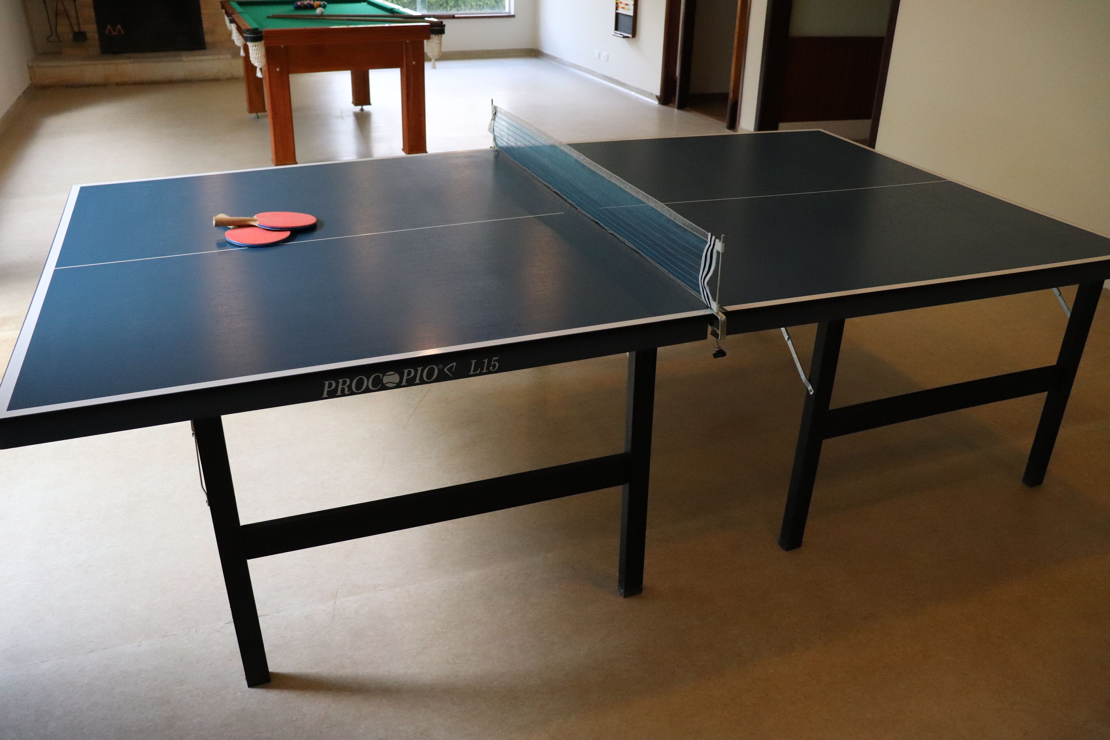 A ping pong table with two paddles in a room with a pool table in the background.