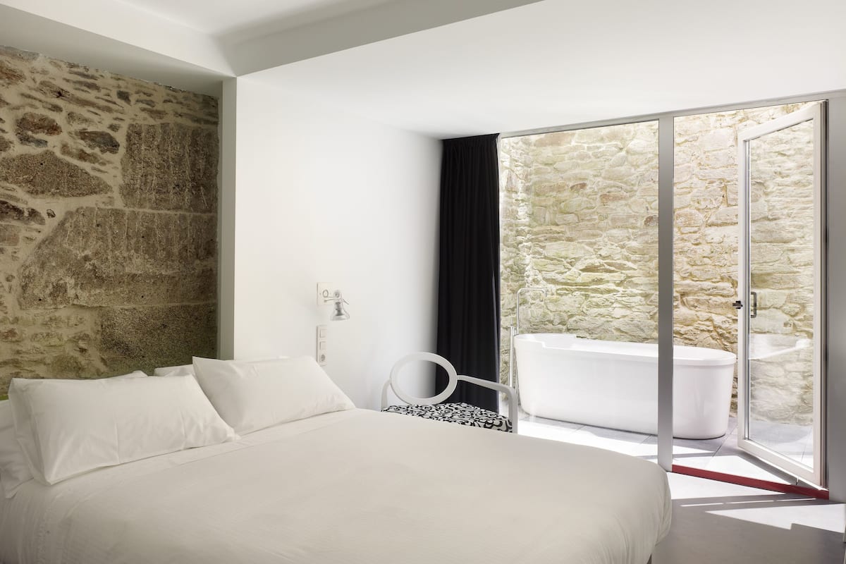 Un dormitorio con pared de piedra, una cama con ropa blanca, una silla blanco y negro y una puerta de cristal que da a un baño con bañera.