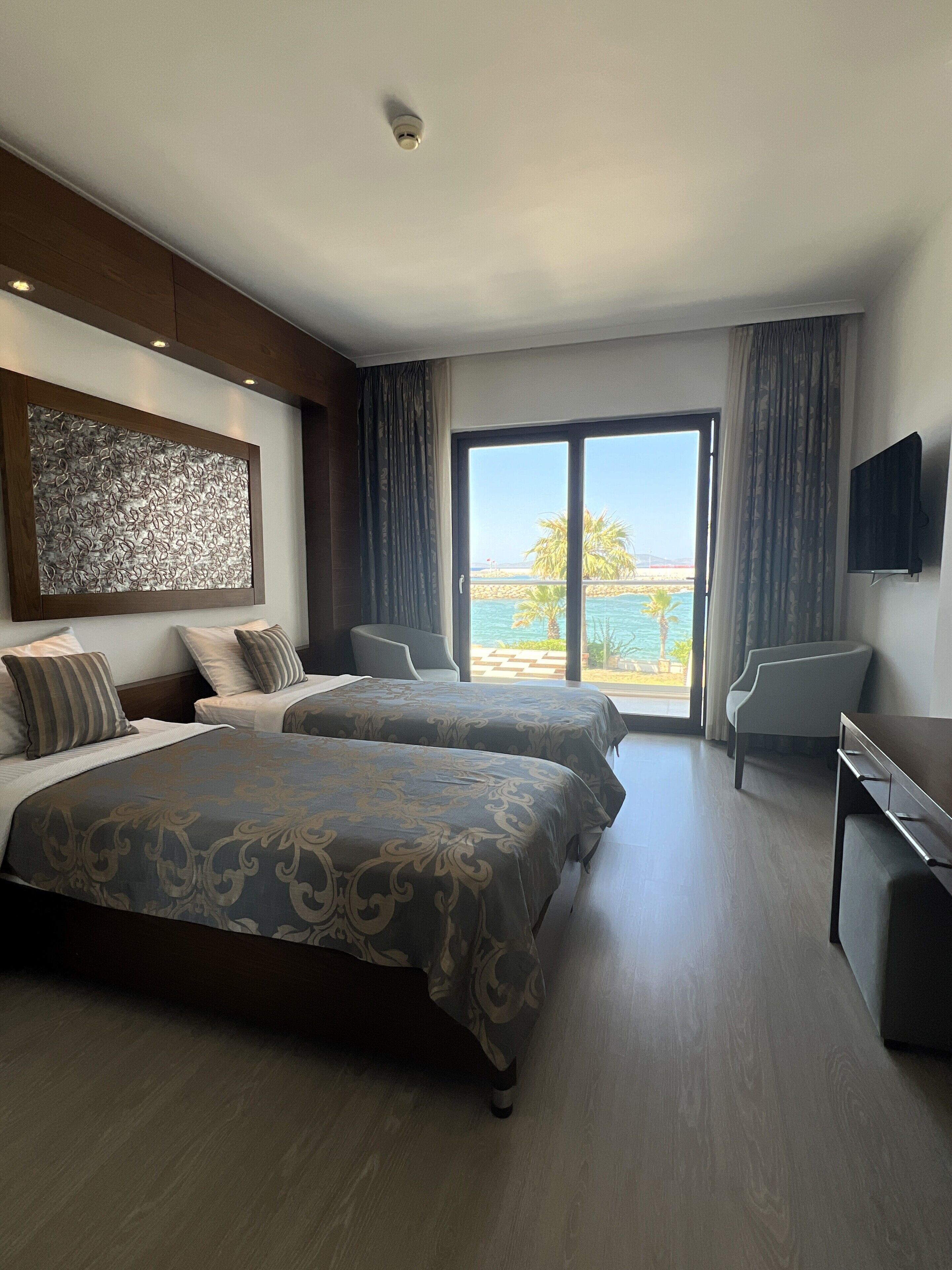 Habitación de hotel con dos camas, televisión, escritorio y balcón con vistas al mar.