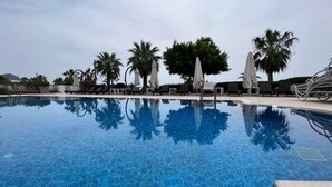 Une piscine aux eaux cristallines, bordée de chaises longues et d’ombrelles, entourée de palmiers et surplombée d’un ciel dégagé.