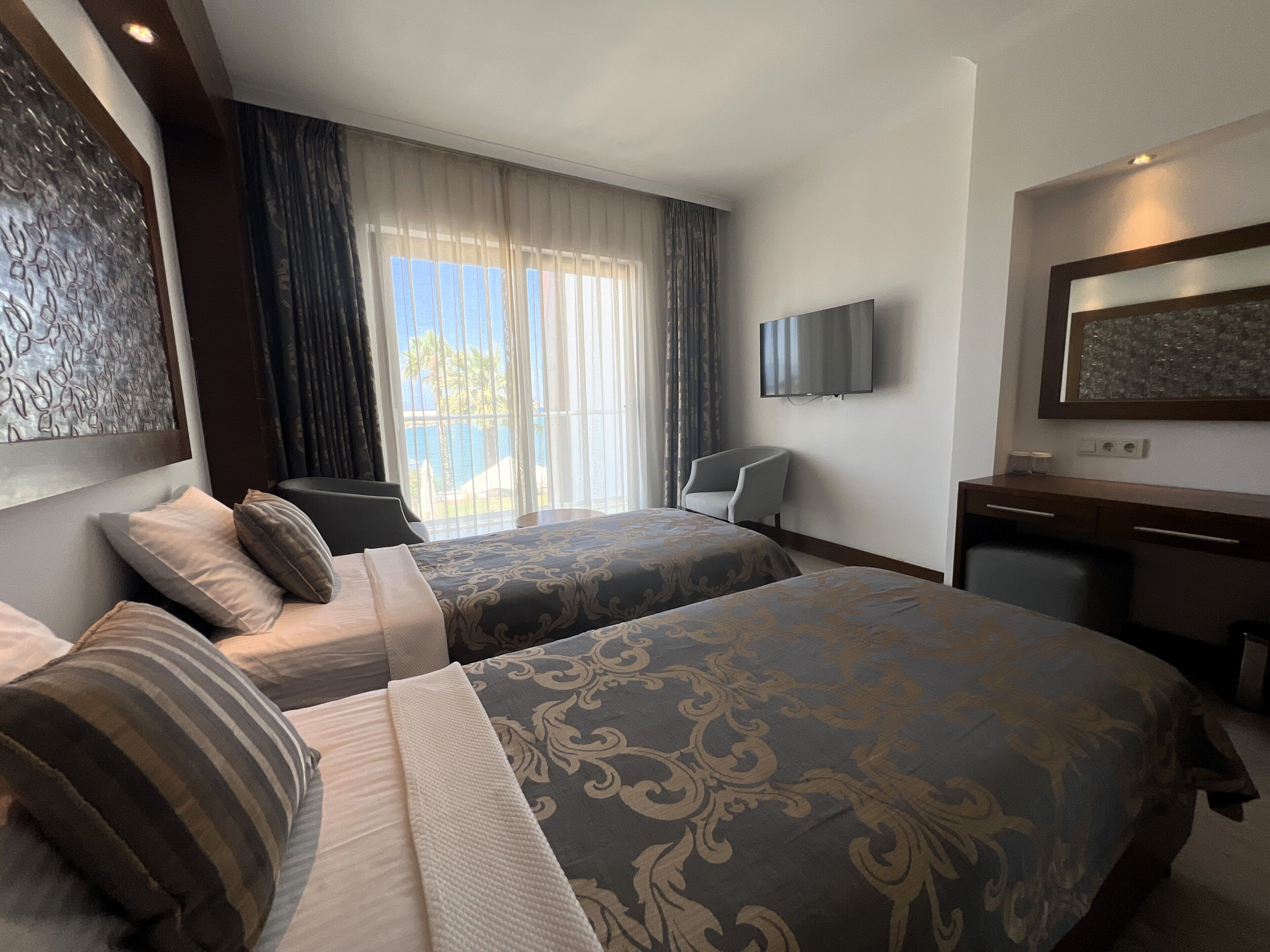 A hotel room with two beds, a desk, a chair, a TV, and a view of the outdoors.