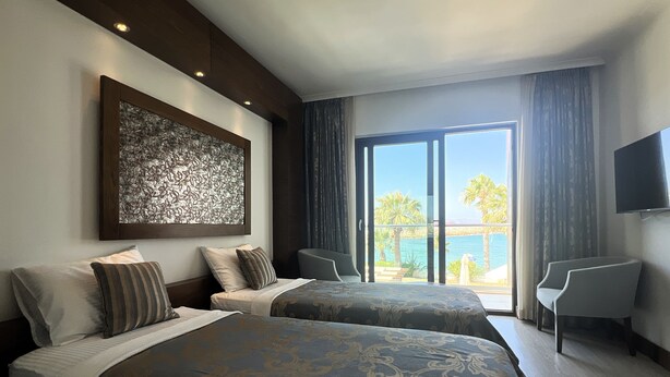 A hotel room with two beds, a large artwork on the wall, a balcony with a view, and a TV mounted on the wall. - Veltur Turiya Hotel (Bodrum)