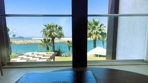 A room with a large window overlooking a beach with palm trees and a white umbrella. - Veltur Turiya Hotel (Bodrum)