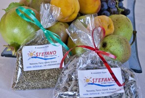 Two gift bags with labels from Stefano, containing dried herbs, placed in front of a variety of fruits. - Stefano Apartments (Paros)