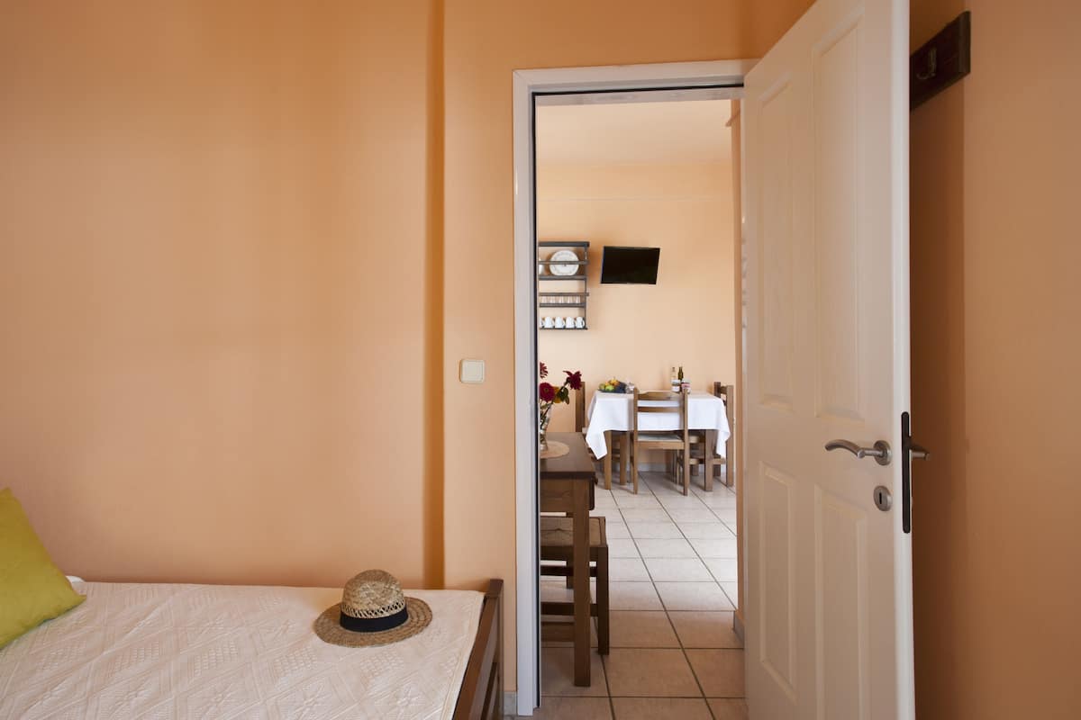 A bedroom with a bed, a hat on the bed, and a door leading to a dining area with a table and chairs.