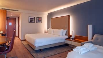 A hotel room with a large bed, a desk, a chair, and a bedside table with a lamp.