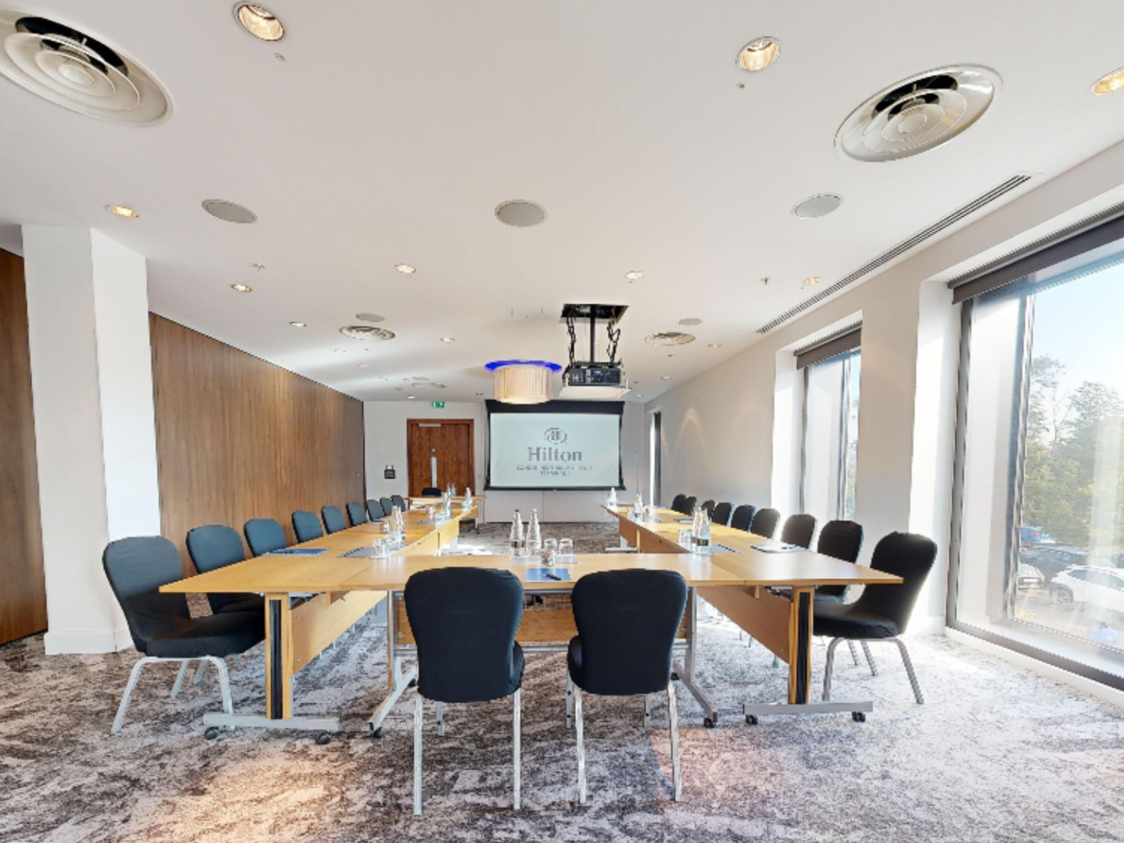 Un salón de conferencias con una mesa larga, sillas, una pantalla de proyector y un ventanal amplio.