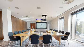 Un salón de conferencias con una mesa larga, sillas, una pantalla de proyector y un ventanal amplio.