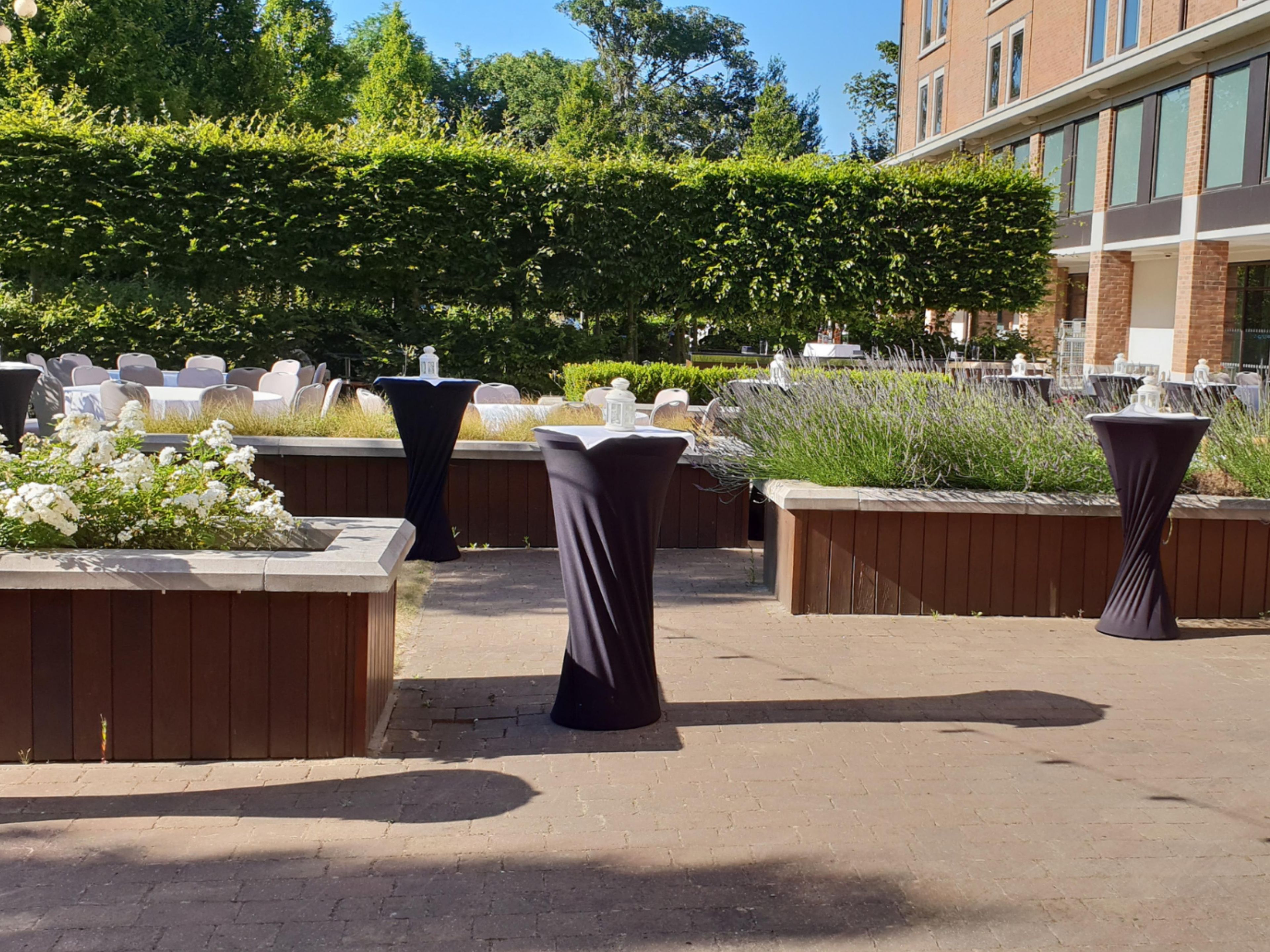 An outdoor seating area with tables and chairs, surrounded by greenery and a building in the background.