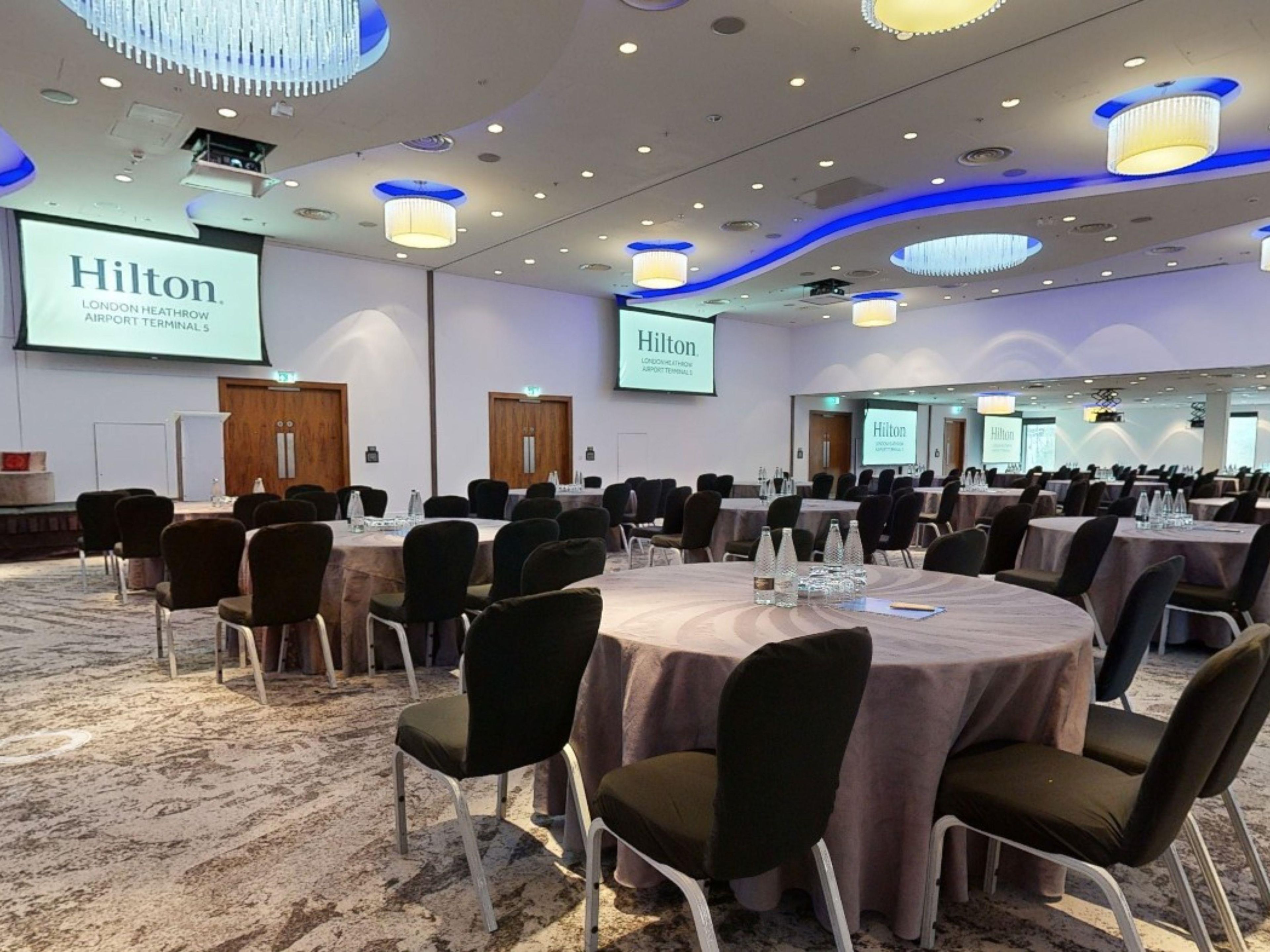 Ein Konferenzraum mit runden Tischen, Stühlen und einem großen Bildschirm, der 'Hilton London Heathrow Airport Terminal 5' anzeigt.