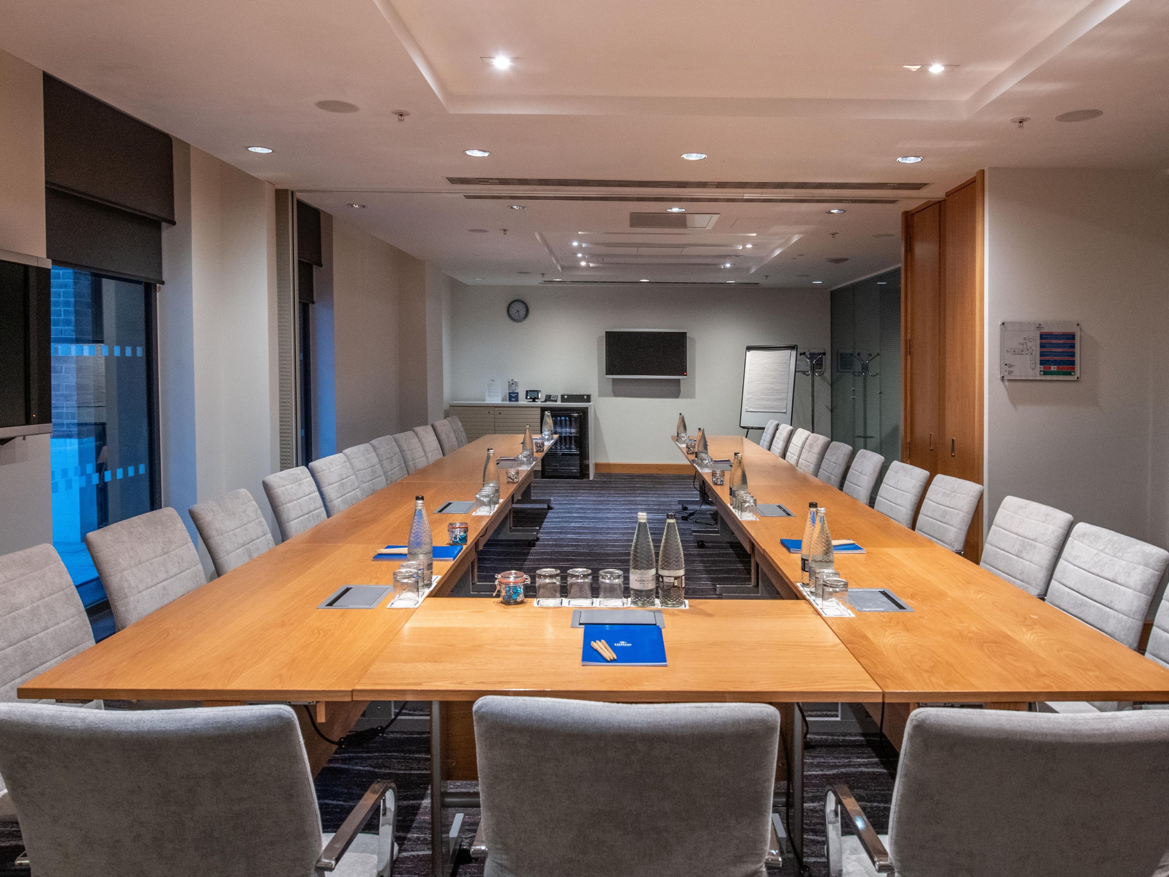 Une salle de conférence avec une longue table, des chaises grises, une horloge, un tableau blanc et un téléviseur fixé au mur.