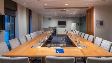 Une salle de conférence avec une longue table, des chaises grises, une horloge, un tableau blanc et un téléviseur fixé au mur.