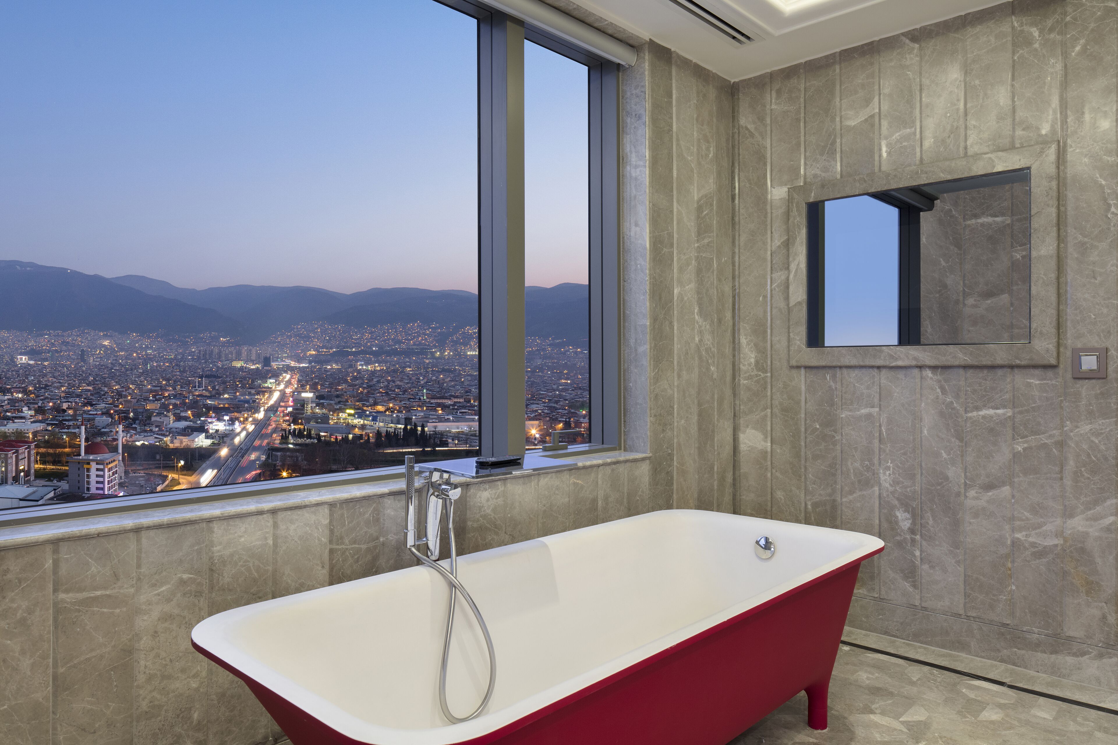 A modern bathroom with a freestanding bathtub, a large window offering a cityscape view, and a marble wall.