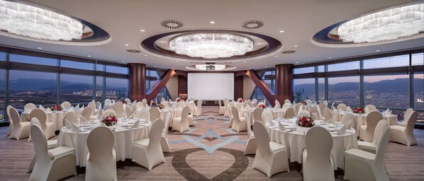 Un salón de conferencias con mesas redondas, sillas blancas y una pantalla grande.