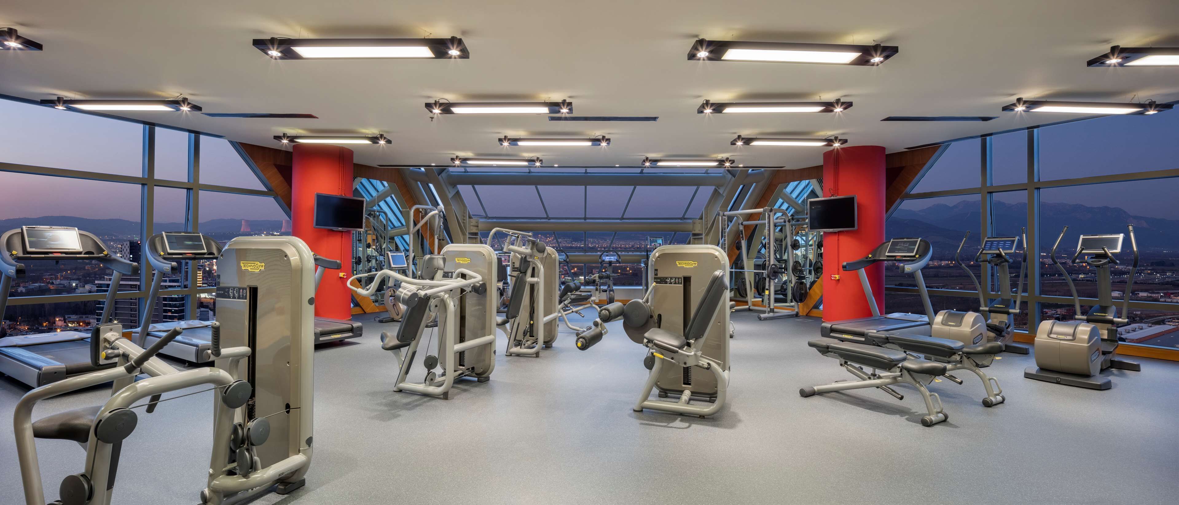 Un gimnasio con diferentes máquinas de ejercicio, grandes ventanales y vista a la ciudad.