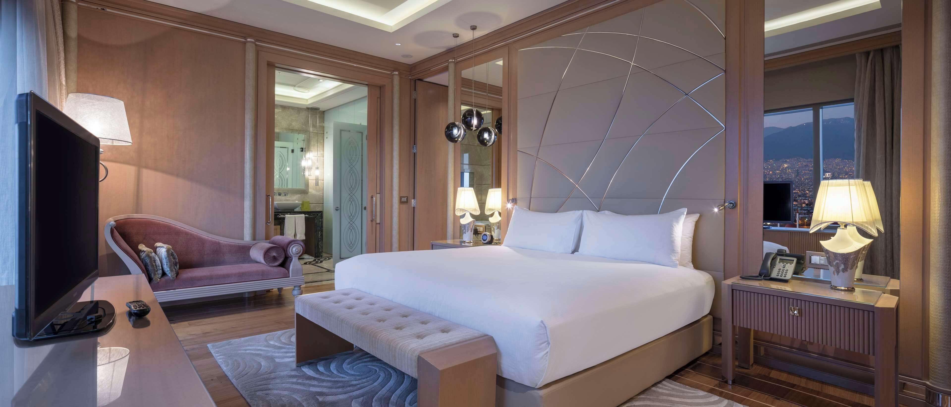 A hotel room with a large bed, a TV, a desk with a lamp, and a view of a cityscape.