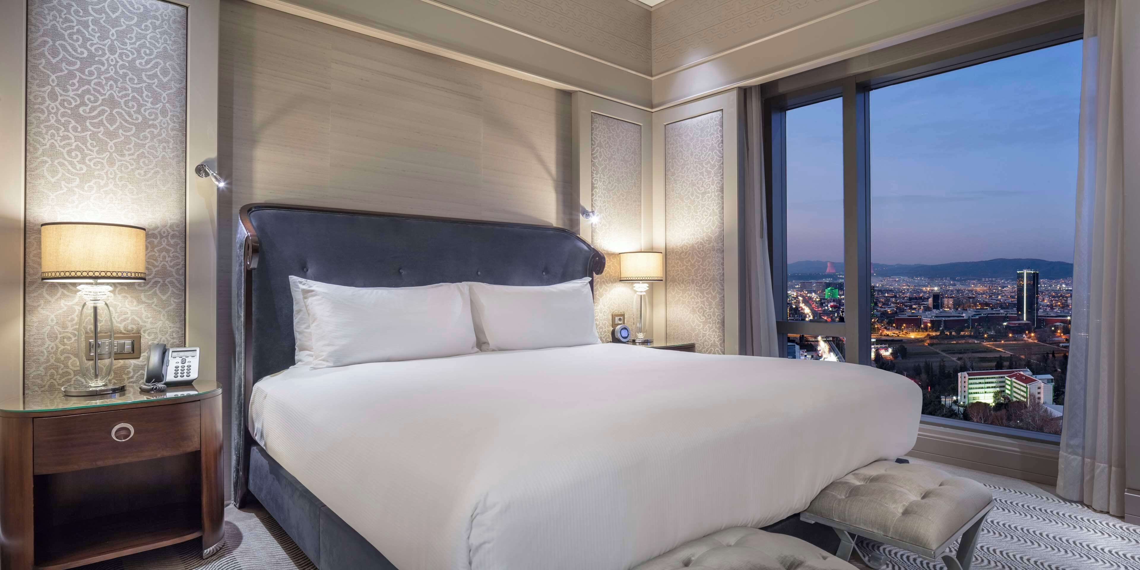 Ein großes Bett mit dunklem Kopfteil, zwei Nachttischlampen, ein Telefon und Blick auf eine Stadtkulisse durch ein großes Fenster.