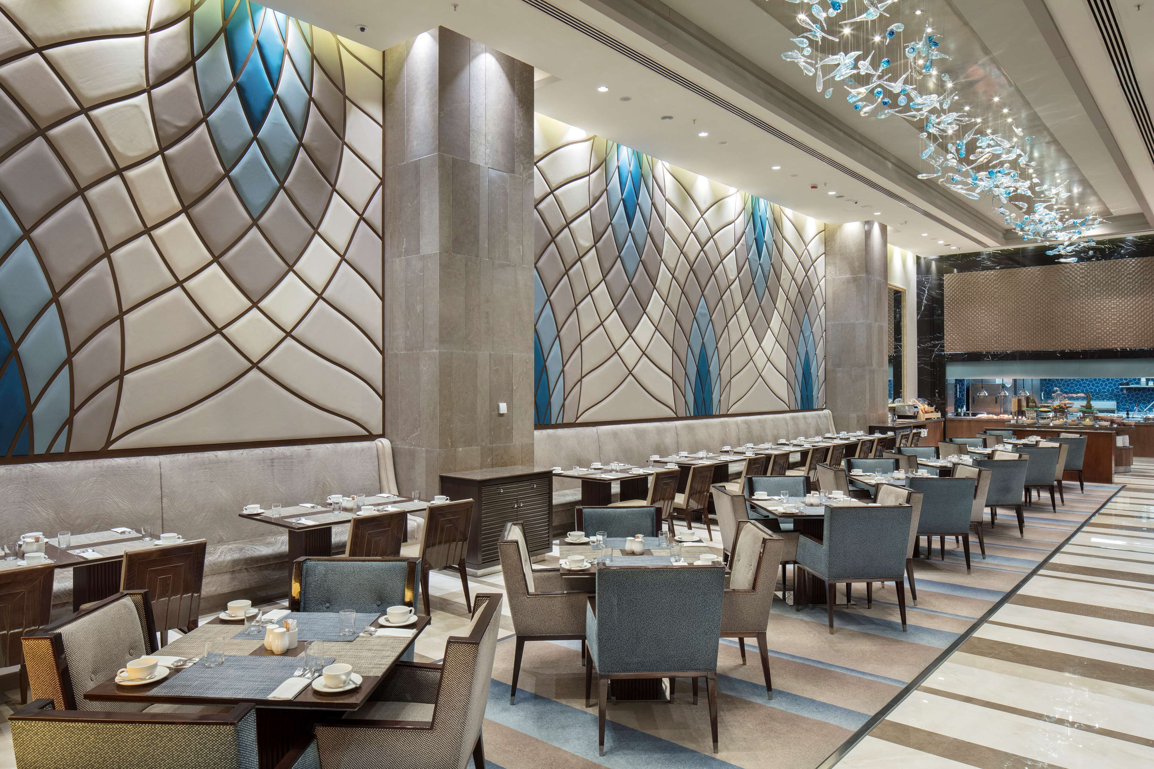 Un espace de restauration moderne, doté de plusieurs tables et chaises, d’un motif décoratif sur un mur et d’un plafond agrémenté de luminaires suspendus.